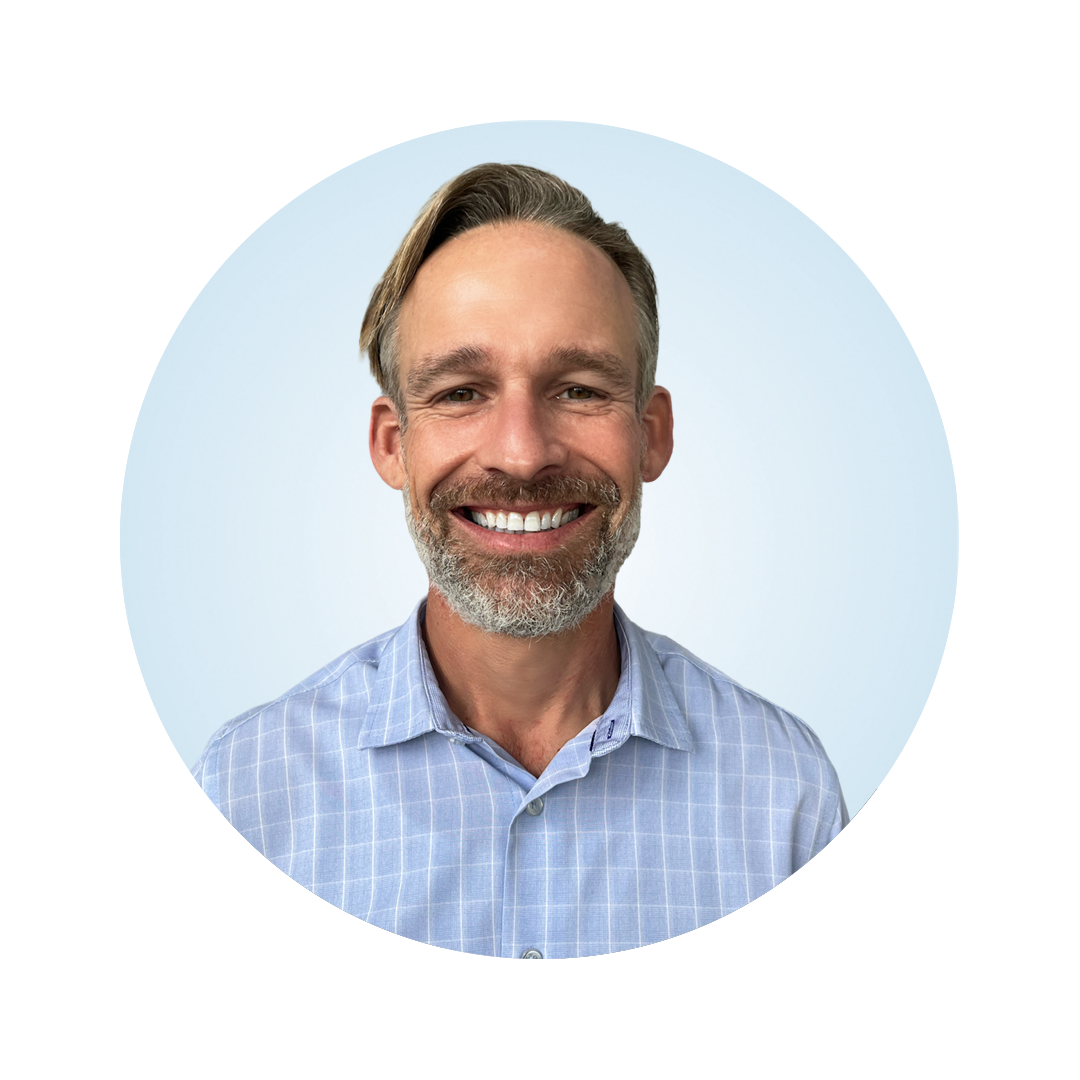 Professional headshot of a smiling man with short hair and a beard, wearing a light blue button-down shirt against a light blue circular background.