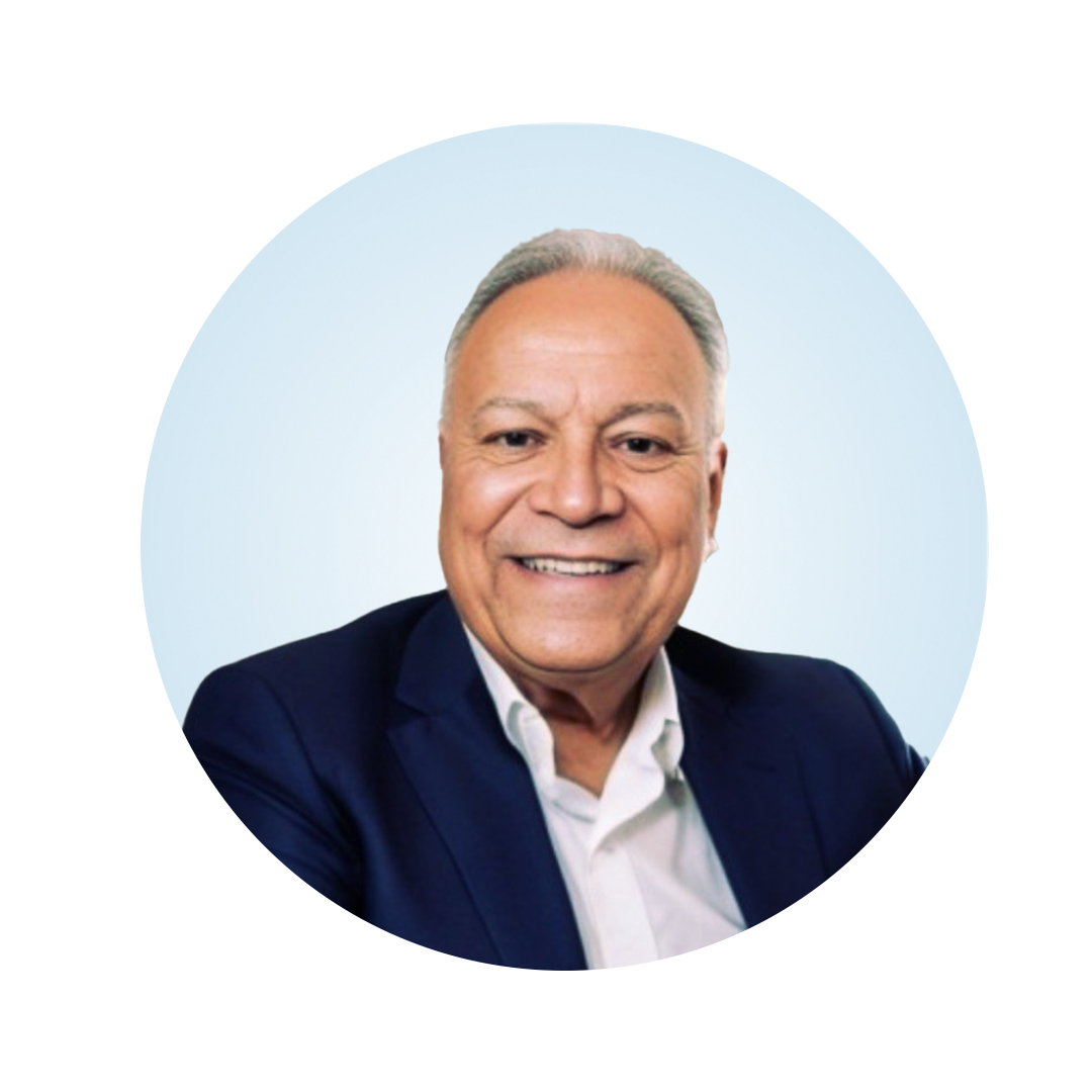 Head-and-shoulders portrait of a smiling older man with short gray hair, wearing a dark blazer over a white collared shirt, against a light blue circular background.