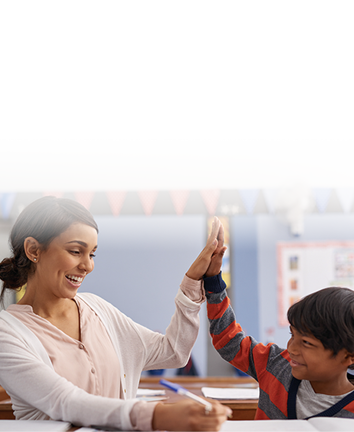 Teacher and elementary student smiling and giving a high-five at a desk in a classroom, celebrating learning success.