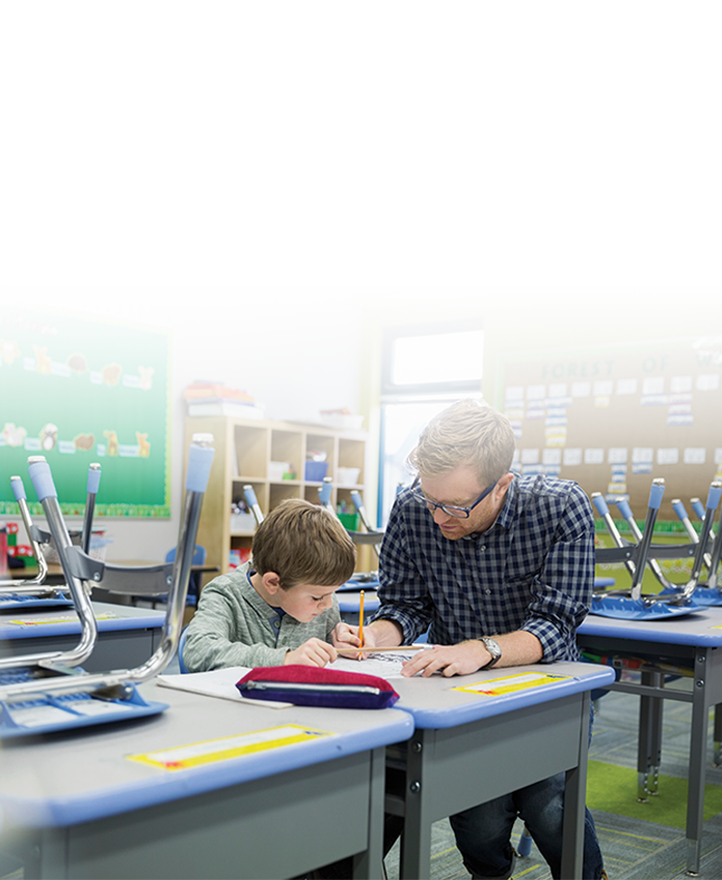 Teacher assisting a young student with classwork at a desk in an elementary classroom, providing one-on-one guidance.